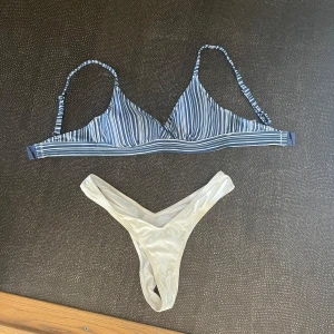Randig bikini med vita trosor - Snygg bikini med blåvitrandig trekants-bh och justerbara axelband. Underdelen är vit och har en v-formad midja med glansigt material och är av stringtrosa-format. Storlek SMALL.