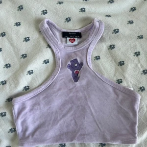 Ljuslila Care Bears linne från Bershka - Supergullig ljuslila linne-topp från Bershka x Care Bears med broderad lila björn framtill. Modellen är croppad och har brottarrygg. Tillverkad i mjuk bomull och polyester, perfekt för en söt och avslappnad stil. Det finns också en rosa på min sida. 