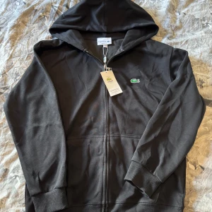 Svart hoodie från Lacoste med dragkedja - Svart hoodie från Lacoste med klassisk krokodillogga på bröstet. Tröjan har huva, hel dragkedja framtill och ribbade muddar. Tillverkad i mjuk bomull och har en regular fit som passar perfekt till en avslappnad stil.