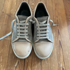 beige sneakers från Lanvin - Snygga sneakers från Lanvin i grå mocka med beige skinn på tå och runt snörningen. Klassisk låg modell med rund tå, platt sula och grå snören. Perfekta för dig som gillar stilrena och neutrala färger med en lyxig touch.