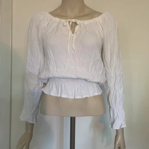 Vit blus med knyt från Hennes - Supersöt vit blus från Hennes med långa ärmar, resår och volang i midjan samt knytning vid halsen. Luftigt tyg och avslappnad passform, perfekt till jeans eller kjol. Offshoulder-vibe och romantisk känsla.