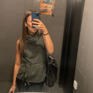  ärmlös topp från Zara - Snygg mörkgrå ärmlös topp med hög krage och knyt i midjan. Perfekt för dig som gillar en stilren look med lite edge. Toppen har en lös passform och är tillverkad i ett mjukt material som känns skönt mot huden.