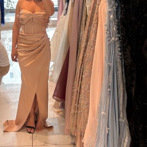 Champagnefärgad långklänning med slits - Supersnygg axelbandslös långklänning i champagnefärgad satin med draperad midja och markerad byst. Klänningen har en elegant slits framtill och en figurnära passform som ger en lyxig känsla.