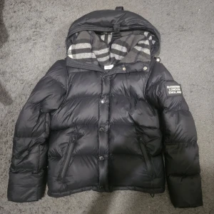 Svart pufferjacka från Burberry - Säljer en snygg svart pufferjacka från Burberry med klassiskt rutigt foder. Jackan har dragkedja och knappar framtill samt en stor logotyp på ärmen. Kan även göras till väst som passar perfekt för höst och vår. Den är storlek S men passar Xs också.