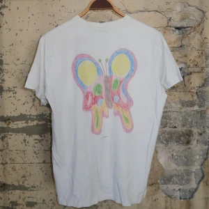 Vit Ganni t-shirt med fjärilstryck - Vit t-shirt från Ganni med tryck av färgglad fjäril på ryggen i pastellfärger och svart logga framtill. Klassisk rund hals och korta ärmar. Skön och avslappnad passform, perfekt till jeans eller shorts. Tillverkad i mjuk bomull.