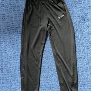 Svarta Nike Dri-Fit träningsbyxor XL (158-170) - Svarta träningsbyxor från Nike med Dri-Fit-teknologi och elastisk midja. Byxorna har en smal passform och är tillverkade i polyester. Swoosh-logga på låret ger en sportig vibe. Perfekta för träning eller chill dagar.