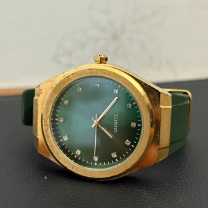 Grön och guldig Quartz klocka - Stilren klocka med grön urtavla och matchande grönt armband i syntet. Boetten är guldig och har en kantig form, medan urtavlan pryds av små glittriga stenar vid varje timmarkering. Perfekt accessoar för dig som vill sticka ut med färg och bling.