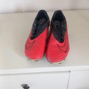Röda Nike fotbollsskor med dobbar - Säljer ett par snygga fotbollsskor från Nike i rött och svart med stora Swoosh-loggor på sidorna. Skorna har snörning och dobbar för bra grepp på planen. Perfekta för dig som vill sticka ut på matchen!