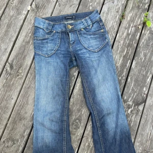 Low waist bootcut jeans - Snygga blå jeans från Lindex i bootcut-modell med coola detaljer som stora fickor fram och bak, samt slitningar för en avslappnad look. Jeansen har klassisk knappstängning och är tillverkade i jeansmaterial med kontrastsömmar.