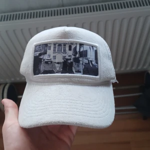 Vit keps från Emilio Pendenza - Vit keps från Emilio Pendenza med mesh på sidorna och baksidan för extra ventilation. Framtill finns en svartvit fotoprint med stadsmotiv. Justerbar snapback baktill och broderad logga på insidan. Perfekt för dig som vill ha en unik och stilren look.