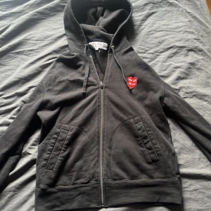 Svart hoodie Comme des Garçons Play M - Svart hoodie från Comme des Garçons Play i storlek M. Klassisk dragkedja framtill, två fickor och huva med snörning. På bröstet sitter det ikoniska röda hjärta-märket med ögon. Mjuk bomullskvalitet, perfekt för en chill look. Passar även storlek S inte använd super mycket