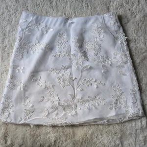 Vit blommig kjol med broderi - Superfin vit kortkjol från bikbok med broderade blommor och blad över hela tyget. Kjolen har en lätt glansig yta och känns riktigt fräsch och somrig. Perfekt för dig som gillar romantiska detaljer och vill sticka ut lite extra.