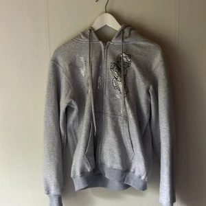 Grå hoodie med ängeltryck Vonstudio - Grå hoodie från Vonstudio i storlek M med ett coolt silverglittrigt ängeltryck på bröstet. Tröjan har huva med snörning och en klassisk känguruficka framtill. Perfekt för dig som gillar streetstyle och vill ha något unikt i garderoben.