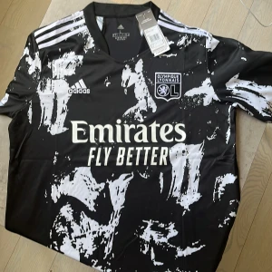 Olympique Lyonnais 21/22 bortatröja, Castello JR #4 oanvänd med prislapp - Snygg Olympique Lyonnais fotbollströja från Adidas i svart med vita abstrakta mönster. Kortärmad modell med klubbmärke, Adidas-logga och Emirates-sponsor på bröstet. Nummer 4 och Castello Jr på ryggen. Tillverkad i lätt och ventilerande AEROREADY-material. Skick 10/10 med prislapp på.