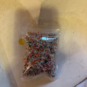 Färgglada pärlor för smyckestillverkning - En påse med massor av små, färgglada pärlor i olika nyanser. Perfekt för att göra egna armband, halsband eller andra kreativa smycken. Pärlorna är runda och kommer i blandade färger som rosa, blå, grön, vit, lila och orange.