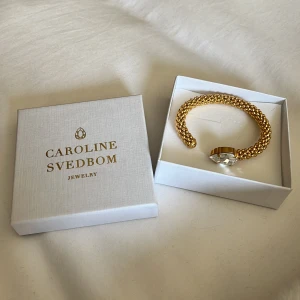 Guldarmband med kristall Caroline Svedbom - Exklusivt armband från Caroline Svedbom i guldton med en stor, droppformad klar kristall. Armbandet har en chunky design med små guldfärgade kulor och är öppet framtill för enkel påtagning. Perfekt statement piece för att lyfta din look. Nypris 995kr. 💕