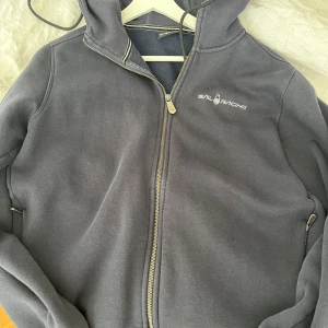 Mörkblå hoodie från Sail Racing - Mörkblå hoodie från Sail Racing med dragkedja och huva. Tröjan har broderad logga på bröstet, snörning i huvan och mjukt fleecefoder på insidan. Perfekt för kyliga dagar och snygg till jeans eller joggers.