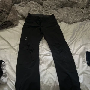 Svarta täckbyxor från Haglöfs - Svarta täckbyxor från Haglöfs med dragkedja och knappstängning i midjan. Byxorna har diskret logga på låret och praktiska fickor med dragkedja. Perfekta för utomhusaktiviteter och gjorda i slitstarkt syntetmaterial.