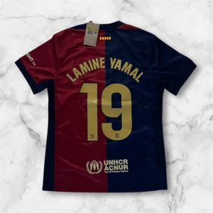 FC Barcelona Lamine Yamal tröja Nike - Snygg FC Barcelona matchtröja från Nike med Lamine Yamal och nummer 19 på ryggen. Tröjan är kortärmad och har klassiska vertikala fält i vinrött och marinblått, med guldiga detaljer och tryck. Materialet är lätt och andas, perfekt för fotboll eller att bära som supporter.