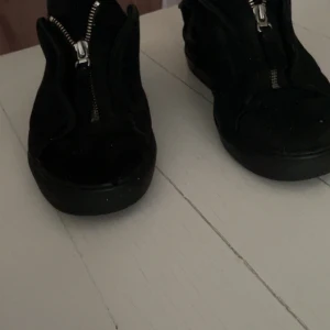 Svarta sneakers med dragkedja - Svarta sneakers med cool dragkedja framtill istället för klassisk snörning. Skorna har rund tå och platt sula, vilket ger en avslappnad vibe. Materialet ser ut att vara mocka eller syntet. Perfekta för dig som gillar en stilren och edgy look. Storleken har töjts ut och passar till storlek 41-41,5.