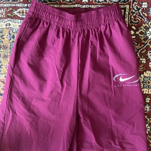 Vinröda Nike byxor - Säljer ett par vinröda byxor från Nike sportswear med elastisk midja. Byxorna har resår vid bensluten och en Nike Sportswear-logga på ena benet.