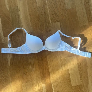 Vit vadderad bh med justerbara band - En enkel och stilren vit bh med vadderade kupor och justerbara axelband. Bh:n har hak- och hyskknäppning bak och en liten rosett mellan kuporna. Materialet är mjukt och stretchigt för extra komfort.