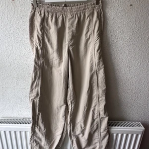 Beiga parachute trousers från Uniqlo - Säljer ett par beiga byxor från Uniqlo med avslappnad passform och resår i midjan. Tillverkade i 100% nylon, perfekta för en chill och bekväm stil. Byxorna har raka ben men resår i botten och praktiska fickor på sidorna. Står storlek L men köpta i thailand så små i storlekarna.