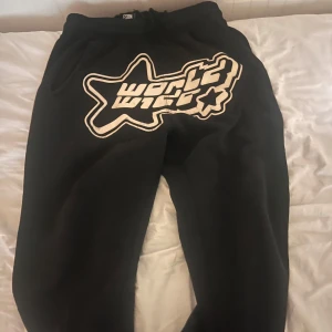 Svarta mjukisbyxor FSBN XS - Svarta mjukisbyxor från FSBN i storlek XS med stor vit grafisk text och stjärndetalj framtill. Byxorna har resår i midjan med snörning och är tillverkade i mjukt material för maximal komfort.