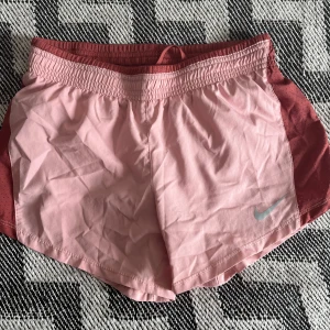 Nike Dri-Fit rosa träningsshorts - Nike Dri-Fit shorts i ljusrosa med mörkare rosa detaljer på sidorna och midjan. De har elastisk midja med snörning och innerficka, perfekt för småsaker. Lätta och snabbtorkande i syntetmaterial, designade för träning och löpning. Shortsen är använda men i fint skick, och i storlek XS.💪