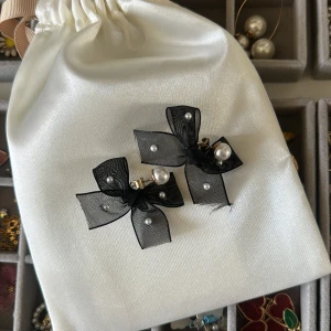 Svarta rosettörhängen med pärlor - Supersöta örhängen med svarta rosetter i tunn mesh och små vita pärlor. Rosetterna är fästa på guldfärgade stift och har en elegant, lekfull vibe. Perfekta för att lyfta vilken outfit som helst med en touch av sötma och stil.