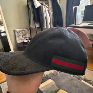 Svart Gucci keps med GG-mönster - Snygg svart keps från Gucci med klassiskt GG-monogram över hela kepsen och ikoniskt grönt och rött band på sidan. Justerbar rem baktill i skinn för perfekt passform. Perfekt accessoar för att lyfta din streetwear-look. 