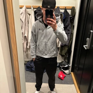 Grå zip hoddie från Polo Ralph Lauren med mörk blått polo märke  - Säljer en grå zip hoddie från Polo Ralph Lauren med dragkedja och huva. Tröjan har mjuk fleece på insidan, har blått polo märke vid bröstet och är i storleken 160-170