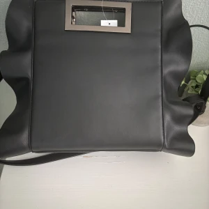 Svart axelväska med snörning och volang - Cool svart axelväska i skinnimitation med vågiga kanter och snörningsdetaljer på sidorna. Väskan har både axelrem och metallhandtag, samt dragkedja inuti. Perfekt för dig som vill ha en unik och edgy accessoar.