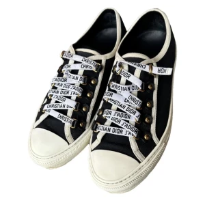 Christian Dior svarta sneakers med logoband - Svarta sneakers från Christian Dior med vita detaljer och coola vita snören med svart logotyptext. Skorna har rund tå, platt sula och mässingsfärgade öljetter. Ovandelen är i canvas och sulan är vit med svart stjärndetalj undertill. Bra skick inga skador, och de är självklart äkta, köpta på Vestiaire 