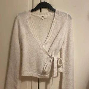 Vit omlottkofta från Gina Tricot - Supermjuk vit omlottkofta från Gina Tricot i ullblandning. Koftan har långa ärmar, ribbade kanter och knytband i sidan för en snygg passform. Perfekt att slänga på sig över ett linne eller t-shirt för en chill och stilren look.
