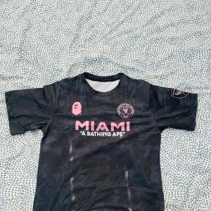 Inter Miami x BAPE fotbollströja svart - Snygg svart Inter Miami fotbollströja i samarbete med BAPE. Tröjan har rosa tryck med MIAMI och BAPE-logga på bröstet samt klubbmärke. Kortärmad modell i lätt och sportigt material, diskret camo-mönster i tyget och tryck på ena ärmen.