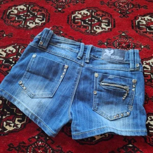 Blå jeansshorts från Monday Supreme - Snygga blå jeansshorts från Monday Supreme med coola detaljer som nitar, dekorativa knappar och dragkedjor på fickorna. Low waisted och perfecta till sommaren. Pris kan diskuteras!