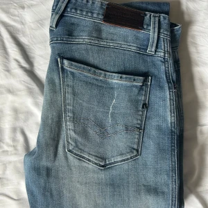 Replay Anbass Jeans Ljusblå - Snygga ljusblå jeans från Replay i storlek W33 och L32, passar även W32, jeansen har slitna detaljer och distressed look. Modellen har klassisk femficksdesign. Jeansen är i mjukt denimtyg och har Replay-logga på bakfickan. Perfekta för en avslappnad streetstil.
