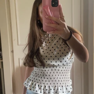 Vit prickig blus med smock - Supersöt vit blus med svarta prickar och smockad midja som ger en snygg form. Toppen är ärmlös och har volangkant nertill för en extra gullig touch. Perfekt till jeans eller kjol för en fräsch look.