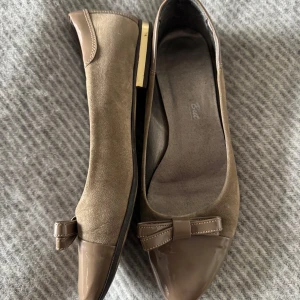 Spetsiga ballerina - Snygga beige ballerinaskor från okänt märke med spetsig tå och lackad detalj framtill. Skorna har en söt rosett på tån och är gjorda i mocka med lackade partier. Sitter väldigt fint under ett par byxor! Storlek 38 