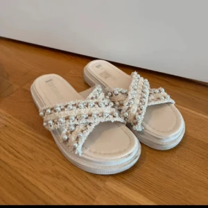 Beige sandaler med pärldetaljer - Snygga beige sandaler från Lemodi med korsade band dekorerade med pärlor och fransiga kanter. Platt sula och öppen tå gör dem perfekta för varma dagar. Ger en trendig och chic vibe till din sommaroutfit.