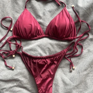 Vinröd bikini  - Snygg vinröd bikini med trekantsöverdel och knyt i sidorna samt runt nacken. Underdelen har höga skärningar och knyt i sidorna med guldiga detaljer på snörena.👙☀️