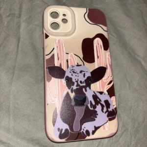 Ko mobilskal IPhone 11 - Unikt mobilskal i beige med bruna och lila detaljer, prytt med en tecknad ko och kaktusar. Skalets design har coola kontraster och sticker verkligen ut. Passar perfekt för dig som gillar djurmotiv och vill ha något annorlunda till mobilen.
