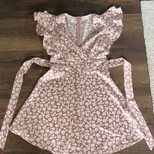 Rosa blommig kortklänning med volang - Supersöt rosa kortklänning med vita blommor, volangärmar och v-ringning. Omlottkänsla med knytband i midjan som ger en snygg siluett. Perfekt för sommarens alla tillfällen och ger en romantisk vibe.