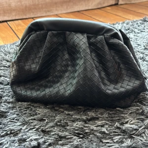 Svart kuvertväska i flätat skinn - Snygg svart kuvertväska med flätad design och mjukt skinnmaterial. Väskan har en stadig ram upptill och avtagbar axelrem för extra flexibilitet. Perfekt storlek för det viktigaste och stängs med metallspänne. Stilren och trendig accessoar.