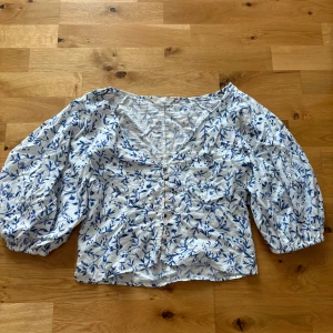 Blommig blus med puffärm och v-ringning - Superfin blus med blått blommönster på vit botten. V-ringad framtill, knappar längs hela framsidan och puffiga långa ärmar med resår vid handlederna. Perfekt för en fräsch och somrig look.