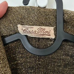 Glittrig guldklänning från Casa Mori - Snygg glittrig guldklänning från Casa Mori med krage, knappar framtill och trekvartsärm. Klänningen har en markerad midja och lätt utställd kjol. Perfekt för dig som vill sticka ut med en elegant och unik look.