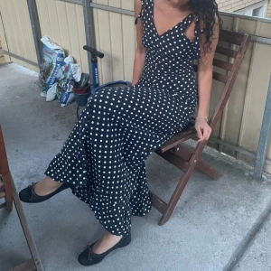 Svart lång prickig klänning med knyt - Supersnygg långklänning i svart med vita prickar och v-ringning köpt secondhand på marknad i Spanien. Klänningen har axelband med knyt och en lätt volang nertill, går ej att justera knyten. Sitter figurnära upptill och faller snyggt längs kroppen. Perfekt för dig som gillar retrovibbar och vill sticka ut. Handsydd, materialet är väldigt hållbart och ganska tjockt, men fortfarande luftigt! Går ner till hälen på mig som är 170