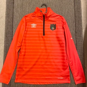 Röd Umbro träningströja - Snygg röd långärmad träningströja från Umbro. Tröjan har diskreta horisontella ränder, halv dragkedja och stårkrage. Tillverkad i snabbtorkande polyester, perfekt för fotbollsträning eller uppvärmning.