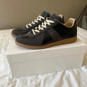 Maison Margiela sneakers svart/grå - Maison Margiela sneakers i svart och mörkgrå mocka och skinn med beige innersida och klassisk gummisula. Snygg retrostil med vita snören och rund tå. Perfekta för dig som gillar minimalistisk och stilren design.